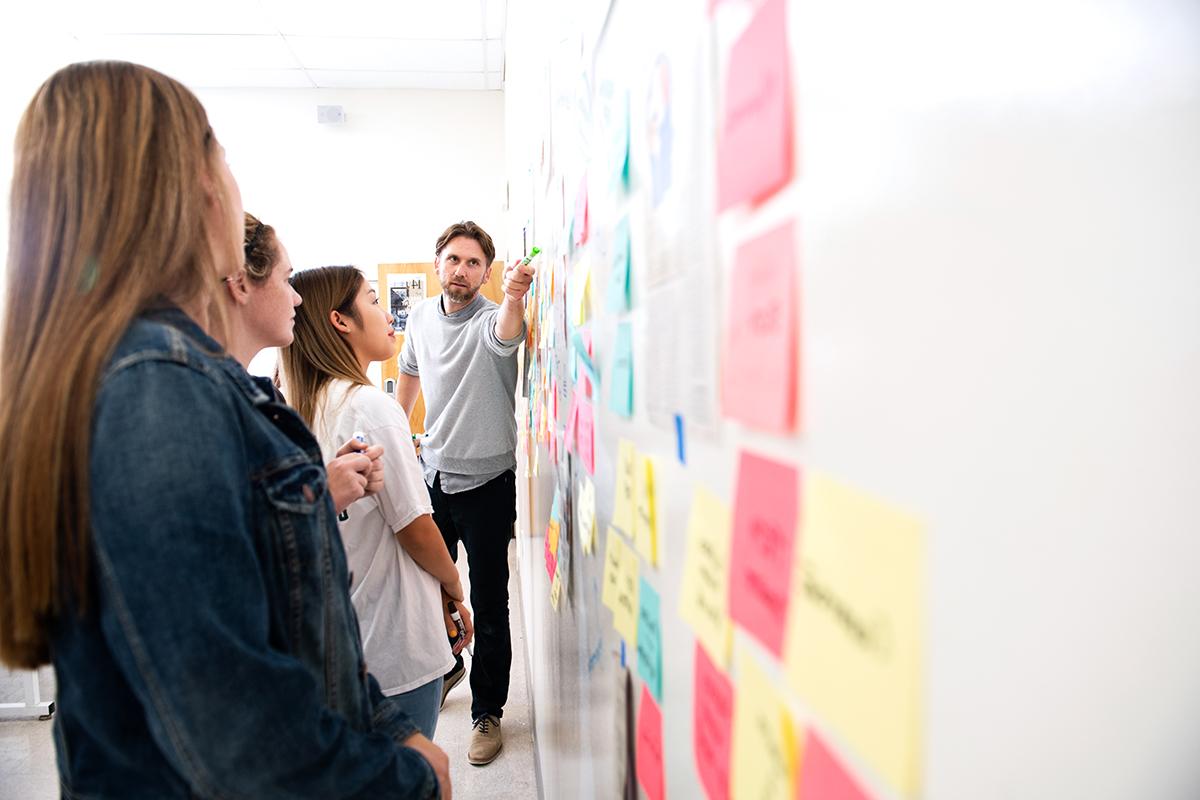 Students and professor engaged in a design thinking exercise using stick notes and collaborating on a writeboard wall.